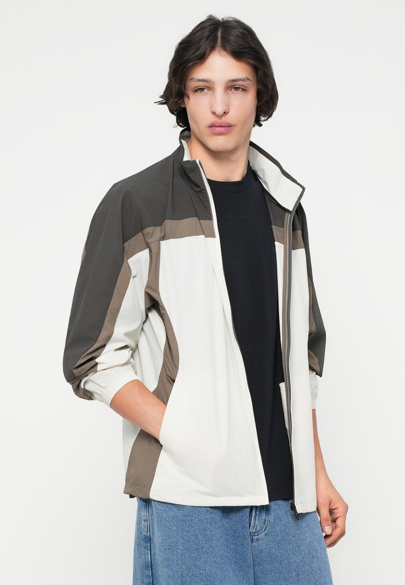 Veste légère bicolore en blanc, marron et gris foncé, avec un col montant, fermeture éclair et poignets élastiques. Matériau synthétique lisse.