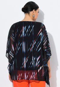 Vloeiende zwarte transparante tuniek met kleurrijk abstract patroon. Heeft wijde mouwen, een high-low zoom en wordt gecombineerd met feloranje broek.