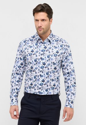 Mand iført hvid skjorte med blå blomsterprint og mørke bukser, stående mod en ensfarvet lys baggrund.