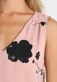 Vaaleanpunainen hihaton toppi, jossa on V-kaula-aukko ja mustia kukkakuviota. Kangas näyttää kevyeltä ja sileäpintaiselta.