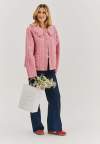 Růžová prošívaná bunda s velkým límcem, knoflíky a kapsami. Modelka má na sobě tmavé kalhoty a červené boty, drží květiny a bílou tašku.