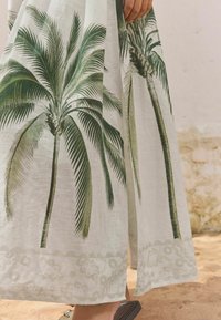 White fabric featuring green palm tree prints with detailed fronds. Hem adorned with a subtle lace-like pattern; lightweight and textured.