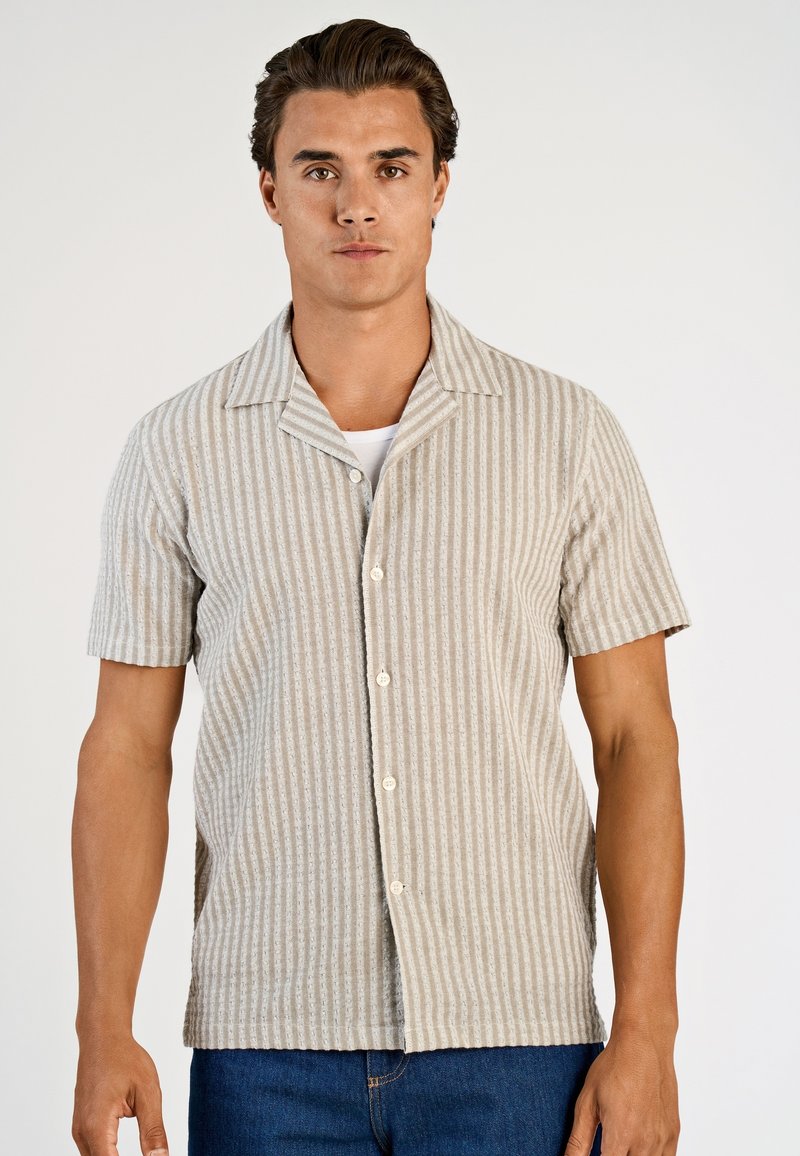 Camicia a maniche corte color beige con righe verticali, realizzata in tessuto strutturato. Presenta un colletto e bottoni frontali.