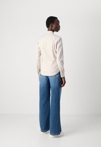 Light beige long-sleeve shirt with a small collar, paired with wide-leg blue denim jeans. Model wears white sneakers, stands against a grey background.