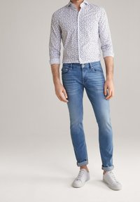Wit shirt met blauw bloemmotief, gecombineerd met lichtblauwe jeans en witte sneakers. Opgerolde mouwen en een slanke pasvorm.