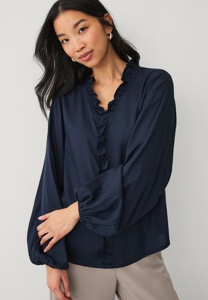 Vrouw met lang donker haar draagt een marineblauwe blouse met gerimpelde kraag en pofmouwen, poseert met gekruiste armen tegen een neutrale achtergrond.