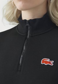 Zbliżenie czarnej bluzki na zamek z wysokim kołnierzem i małym pomarańczowym logo krokodyla po lewej stronie klatki piersiowej, noszonej przez osobę z blond włosami.