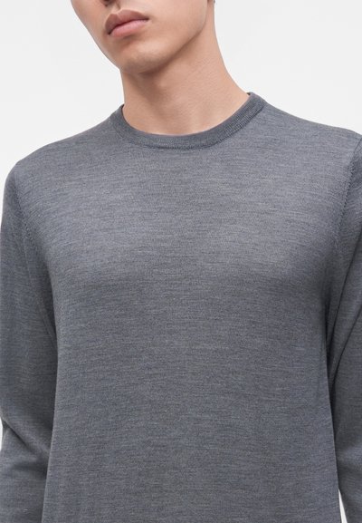Jeune personne portant un pull gris uni à col rond avec manches longues, présenté des épaules jusqu'au milieu de la poitrine sur fond blanc.