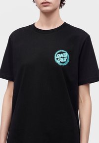 Άτομο φοράει μαύρο T-shirt με μπλε λογότυπο «Santa Cruz» στο αριστερό στήθος, σε απλό φόντο.