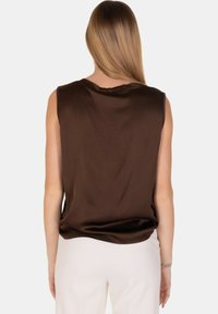 Top senza maniche in satin marrone con una vestibilità rilassata, caratterizzato da una schiena drappeggiata, abbinato a pantaloni bianchi. Texture liscia e design minimalista.