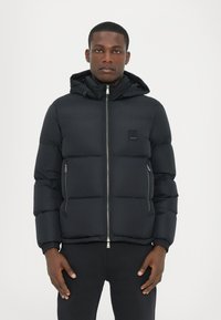 Black puffer jacket with a zip front, hood, and elastic cuffs. Features a textured exterior and a square logo patch on the chest.