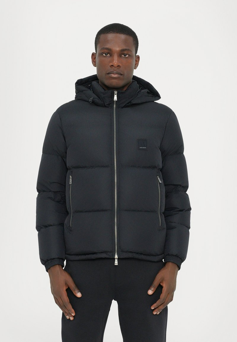 Black puffer jacket with a zip front, hood, and elastic cuffs. Features a textured exterior and a square logo patch on the chest.