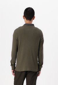 Cămașă polo cu mânecă lungă de culoare verde închis, cu o textură ribbată, guler gri și o croială lejeră care se termină sub talie.