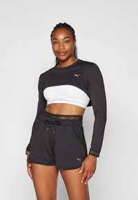 Schwarzes Langarmoberteil mit einem weißen Sport-BH darunter, kombiniert mit schwarzen Shorts mit einem Netzbund. Graues Logo auf dem Oberteil.