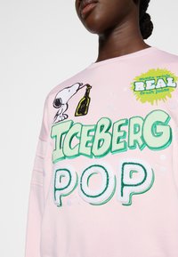 Personne portant un sweat-shirt rose avec Snoopy tenant une bouteille, grand texte vert "ICEBERG POP" et une étiquette jaune indiquant "fabriqué avec du vrai jus de fruit".