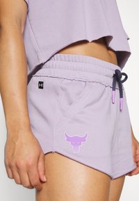 Osoba v levandulových atletických šortkách a odpovídajícím crop topu, s logem býčí lebky na šortkách a stáhnutým šňůrkovým pasem.