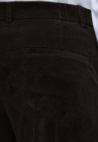 Gros plan sur un pantalon en velours côtelé brun foncé montrant la poche arrière et les passants de ceinture sur fond de chemise blanche.