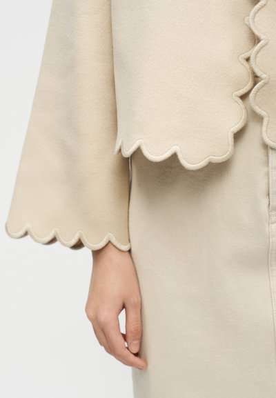 Μανίκι και τελείωμα ρούχου από μπεζ μάλλινο ύφασμα με κυματιστά άκρα, δίπλα σε μπεζ φούστα με ορατή ραφή και χαλαρό χέρι στο πλάι.