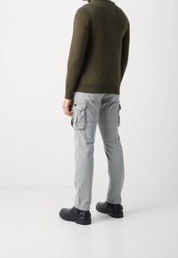 Pull en maille vert olive à manches longues, pantalon cargo gris clair avec poches latérales, bottines noires à boucles, texture lisse, coupe décontractée.