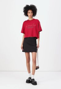 Rotes, kurzes T-Shirt mit "Coach" in Schreibschrift, kombiniert mit einem schwarzen Rock mit Seitenschlitzen. Das Model trägt weiße Socken und schwarze Schuhe.