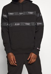 Svart hoodie i mjukt material, med grå "TOMMY JEANS" logostripes över bröstet. Den har en framficka och dragsko i huvan.