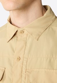 Camicia button-up beige con colletto a punta, caratterizzata da cuciture verticali e due tasche sul petto, realizzata in un tessuto leggero e testurizzato.