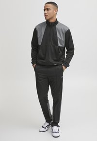 Svart og grå treningsdress med glidelåsjakke og elastiske bukser, med et stilrent design og glatt, lett materiale. Hvite joggesko inkludert.