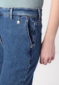 Modre jeans hlače z visokim pasom, z srebrno čarobno obeskom, bakrenimi zakovicami in standardnimi žepi. Gladka tekstura z detajli šivanja.