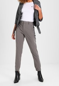 Graue Hosen mit schmalem Schnitt, die eine glatte Textur und Fronttaschen aufweisen, kombiniert mit einem weißen Hemd und einer grauen Lederjacke.