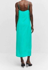 Robe en satin turquoise longue avec des bretelles fines, une silhouette droite et une texture lisse. Portée avec des sandales à talons noires.
