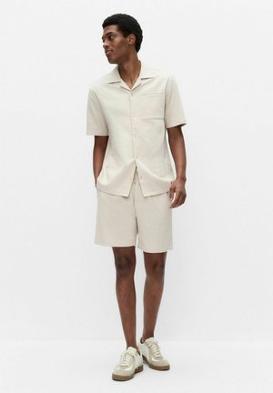 Ung mand stående med hænderne i lommerne, iført off-white kortærmet knap-skjorte, matchende shorts og beige sneakers mod en hvid baggrund.