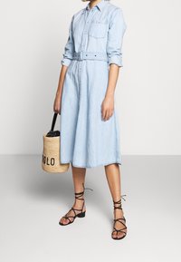 Ljusblå denimskjortklänning med knyten i midjan, knappslå framtill och lång ärm. Svarta sandaler med remmar och en vävd väska med "POLO."