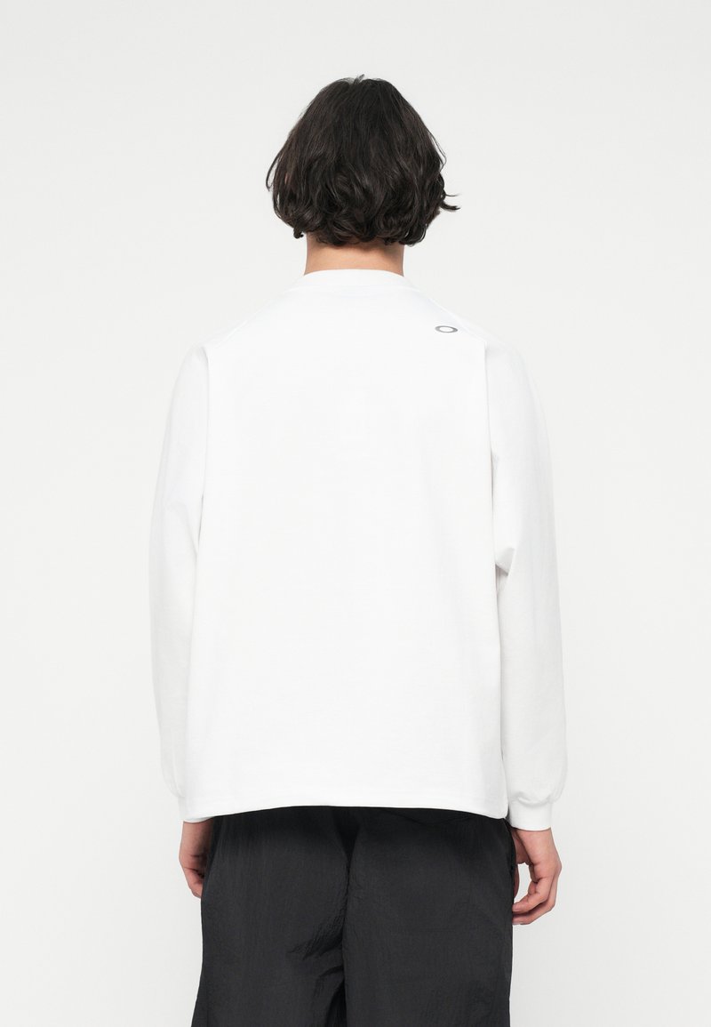 Haut blanc à manches longues avec une coupe décontractée, col rond et un petit logo au dos de l'épaule. Tissu en coton doux, design minimal.