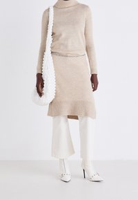 Modèle portant une robe tricot beige à col montant, un pantalon large blanc, des bottes à talons blanches à clous, une ceinture en chaîne argentée et un sac à épaule texturé blanc.