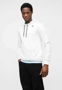 Νεαρός άνδρας που φοράει λευκή μπλούζα με κουκούλα και μαύρα κορδόνια, με μικρό πολύχρωμο λογότυπο, σε συνδυασμό με μαύρο παντελόνι και ανοιχτό μπλε εσώρουχο.