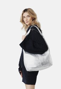 Silverfärgad metallisk tote bag med en mjuk textur, stor form och dubbla axelremmar, hålls av en person i en mörk texturerad tröja.