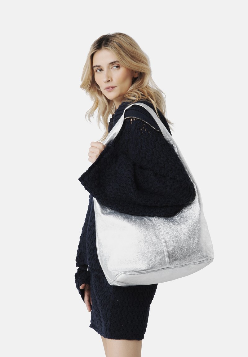 Silverfärgad metallisk tote bag med en mjuk textur, stor form och dubbla axelremmar, hålls av en person i en mörk texturerad tröja.