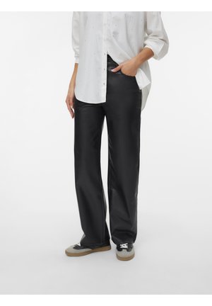 Personne portant une chemise blanche boutonnée avec les manches retroussées, un pantalon noir à jambes larges, et des baskets gris et noir avec des semelles marron.