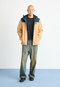 Man with shaved head wears tan and blue jacket over black shirt, loose faded jeans, and black sneakers, standing on blue floor against white wall.