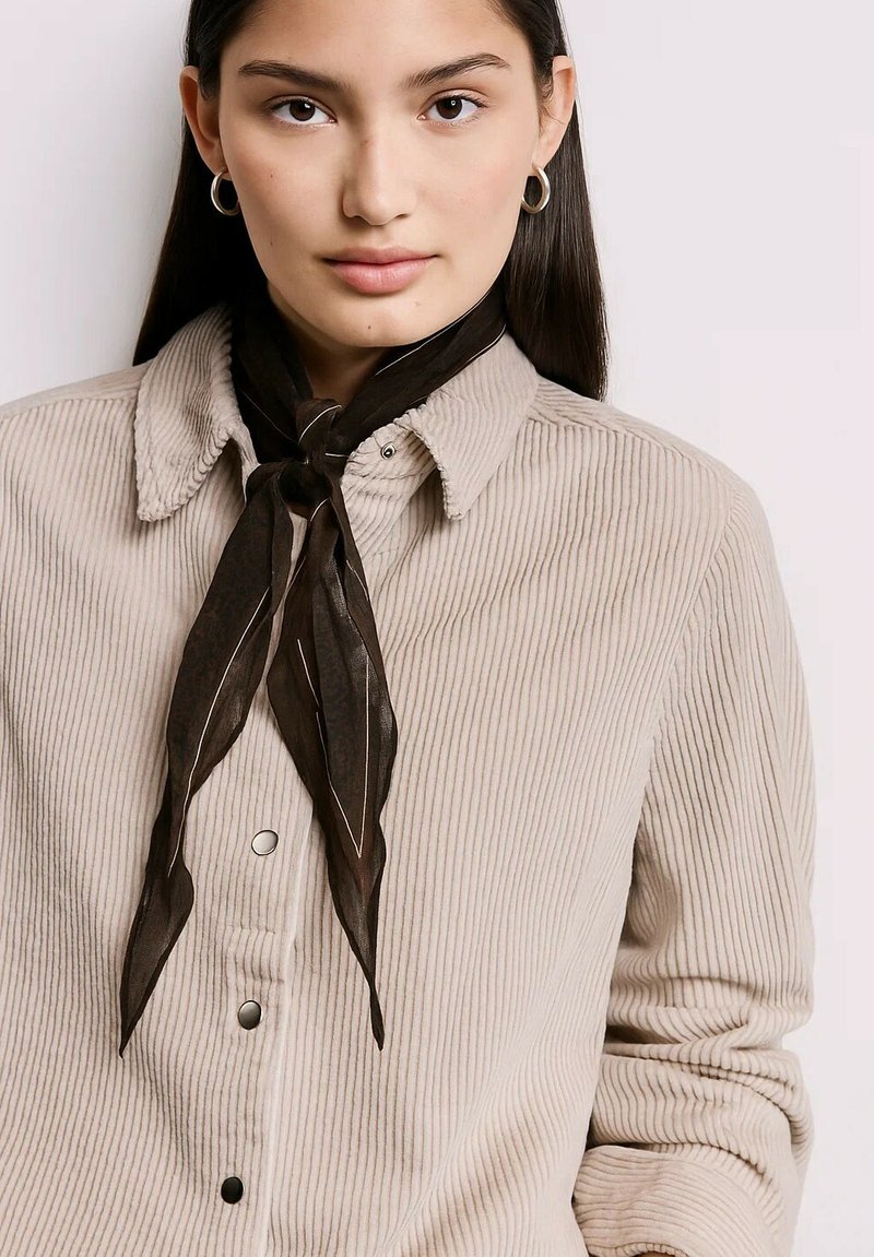 Jeune femme aux longs cheveux noirs portant des boucles d'oreilles en argent, une chemise en velours côtelé beige et une écharpe noire nouée autour du cou.