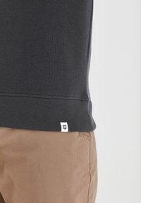 Primer plano de una persona que lleva un sudadera negra con dobladillo acanalado y pantalones marrón claro, mostrando una pequeña etiqueta blanca en el dobladillo de la sudadera.