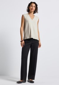 Kvinna som bär en beige ärmlös V-ringad topp, svarta vida byxor, svarta ballerinaskor och ett mörkt armband, står mot en enkel vit bakgrund.