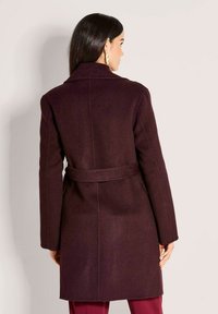 Vrouw met de rug naar ons gekeerd, in een donkerpaarse jas met riem en lange mouwen, en grote gouden oorbellen, tegen een effen lichte achtergrond.