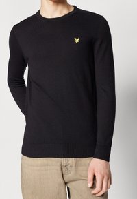Pull en tricot noir à manches longues et col rond. Dispose d'un petit logo jaune sur la poitrine et de poignets et ourlet côtelés.
