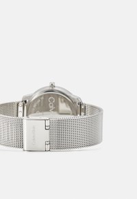 Ura z oglatim ohišjem z srebrno mrežasto zapestnico. Minimalističen dizajn z teksturirano zapestnico in zaponko, označeno z "Calvin Klein."