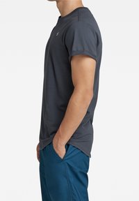 Man i mörkgrå kortärmad skjorta och blåa byxor, stående på sidan med höger hand i fickan mot en enfärgad vit bakgrund.