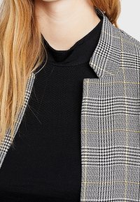 Blazer a cuadros grises con acentos amarillos sobre una blusa negra texturizada. Presenta un cuello de muesca y un diseño de frente abierto.