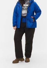 Chaqueta impermeable azul con capucha, suéter gris con estampado, pantalones aislantes negros y botas gruesas marrones. Muestra elementos de diseño funcionales.