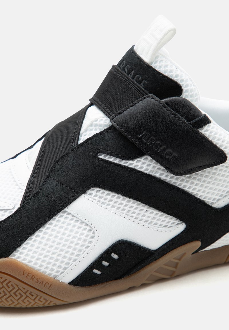 Scarpa sportiva Versace bianca e nera con tessuto in mesh, sovrapposizioni in pelle scamosciata, cinturino in velcro e suola in gomma marrone con motivo a fantasia.