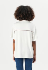 Weißes T-Shirt mit kurzen Ärmeln und strukturiertem Finish, das eine kontrastierende braune horizontale Naht am oberen Rücken aufweist.